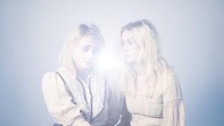 Maustetytöt näkyvät utuisesti valkoisiin paitoihin ja tummiin hameisiin pukeutuneina. Tausta siniharmaa ja keskellä kirkas valo.